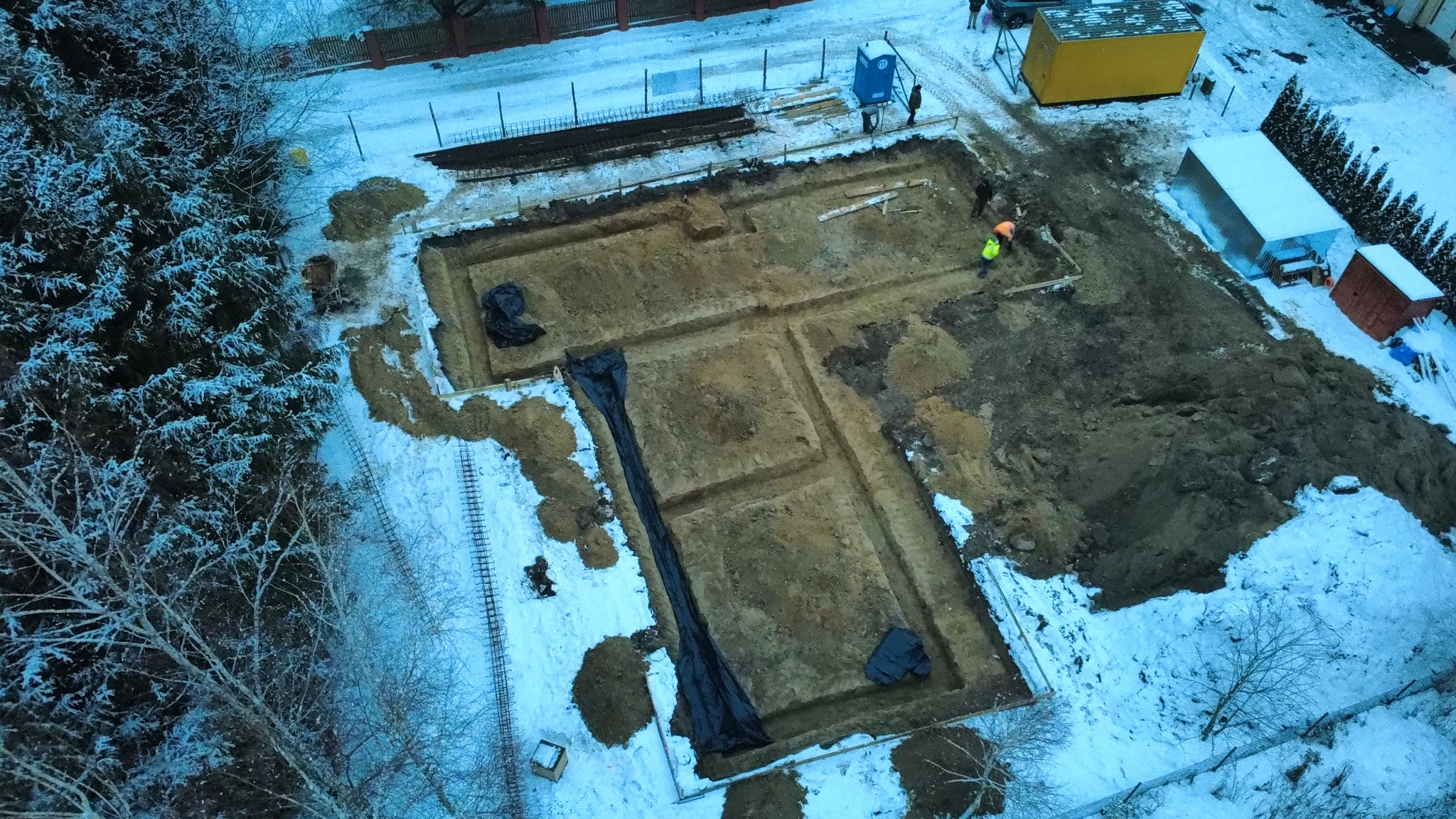 Zimowy widok z góry na wykop pod fundamenty domu, z widocznymi zarysami ścian i pracownikami w odblaskowych kamizelkach. Częściowo ośnieżone otoczenie.