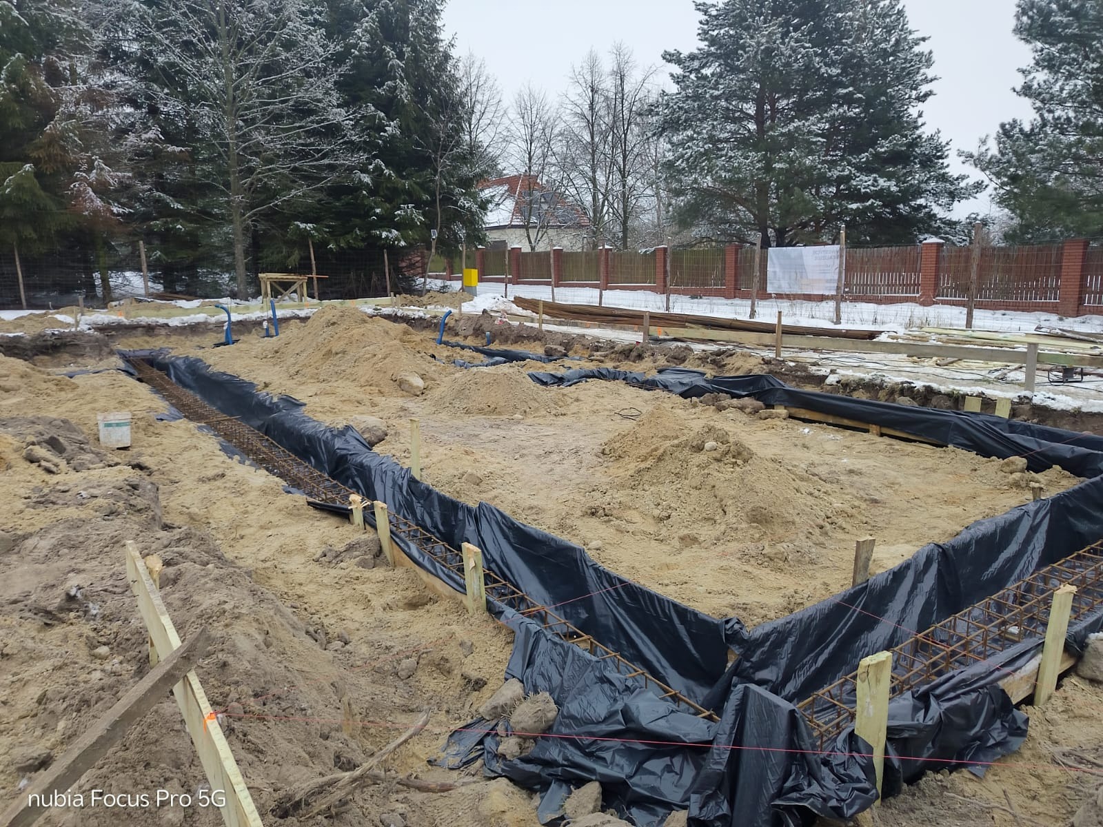 Przygotowanie fundamentów pod dom murowany: wykopane rowy, zbrojenie zabezpieczone folią, drewniane szalunki. W tle drzewa i ogrodzenie.