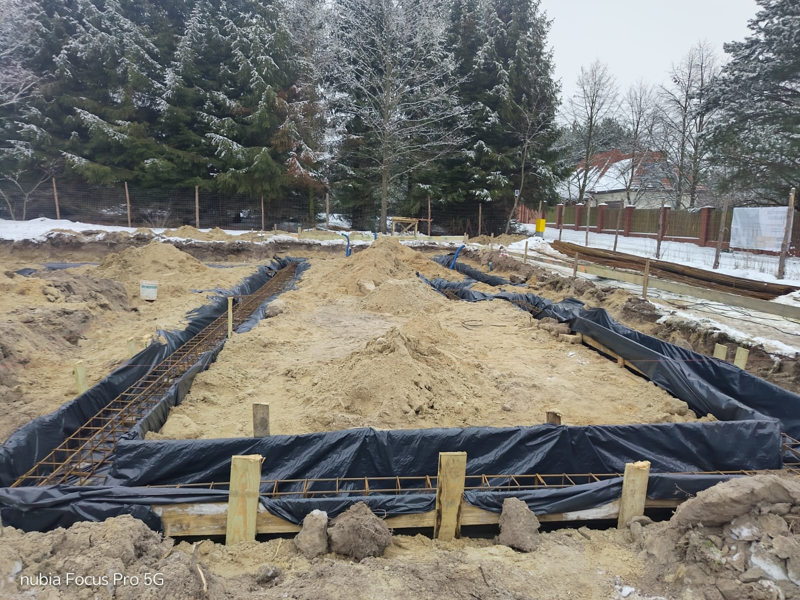Przygotowanie fundamentów pod dom murowany: wykopane rowy, zbrojenie w czarnej folii, drewniane szalunki, ziemia i śnieg w tle.