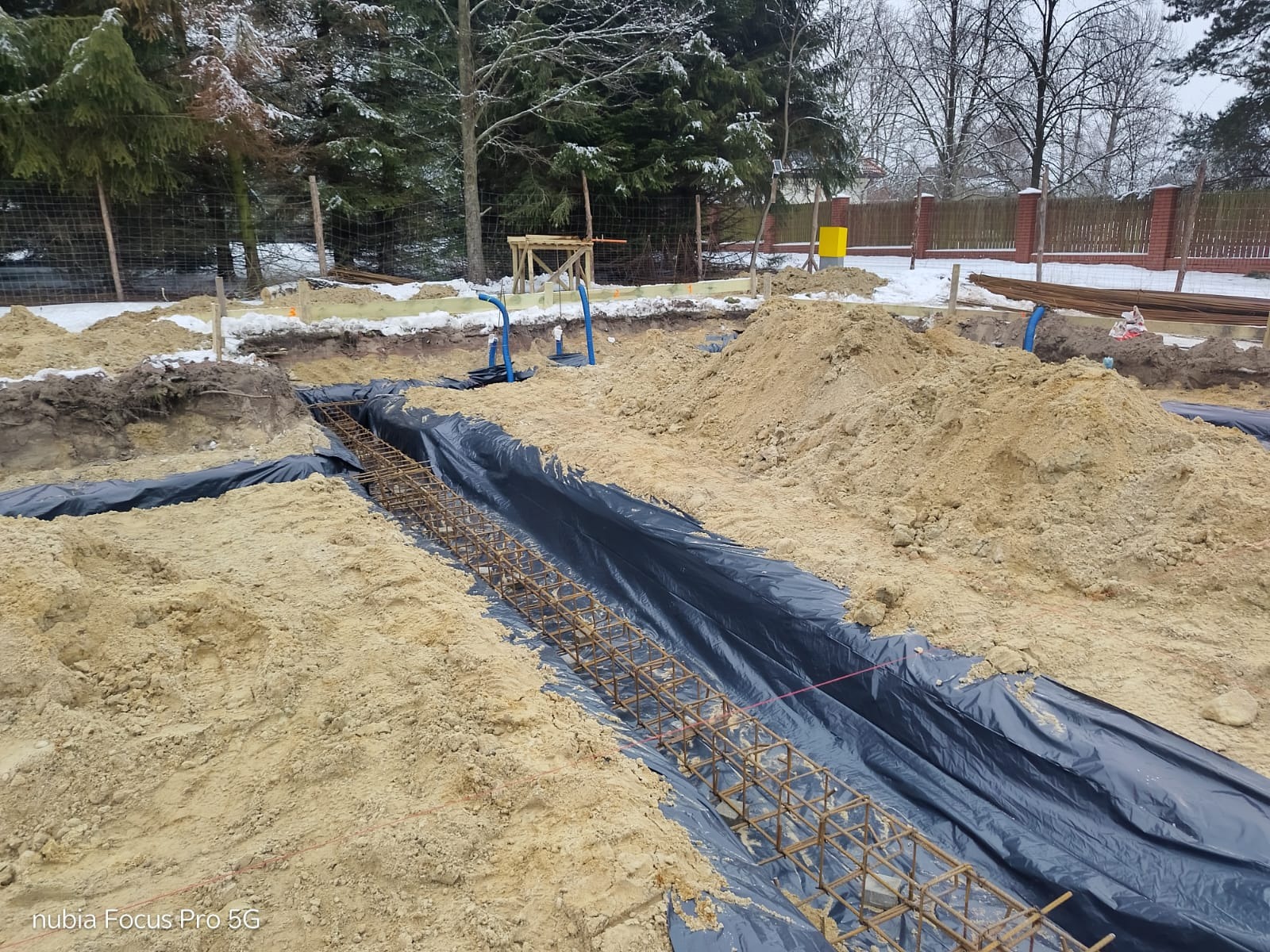 Zbrojenie fundamentów domu jednorodzinnego, zabezpieczone folią, w wykopie. W tle widoczne drzewa i ogrodzenie posesji. Zima, śnieg.