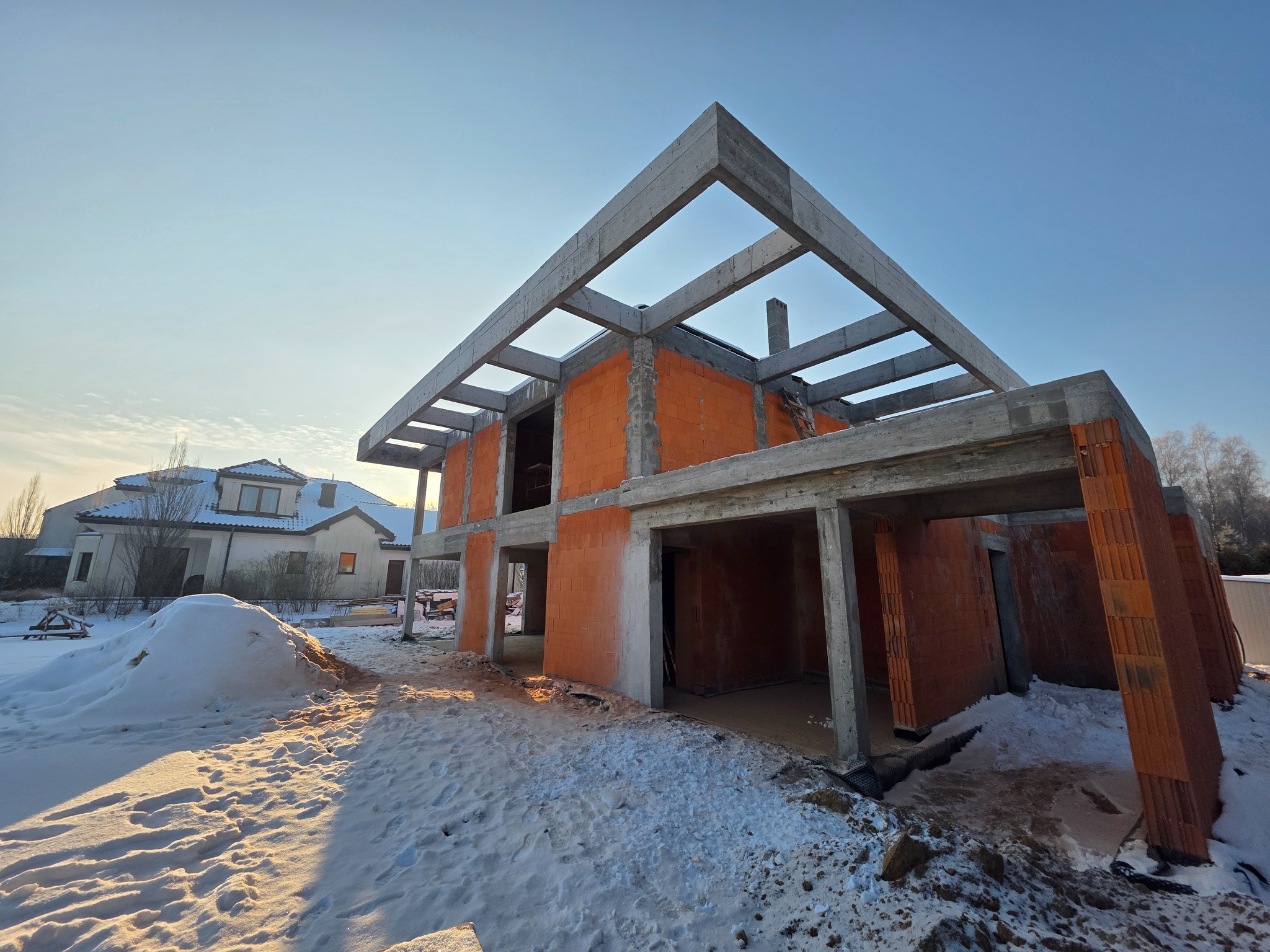 Stan surowy otwarty domu z czerwonej cegły i betonowym szkieletem w zimowej scenerii. W tle widoczny inny dom i błękitne niebo.