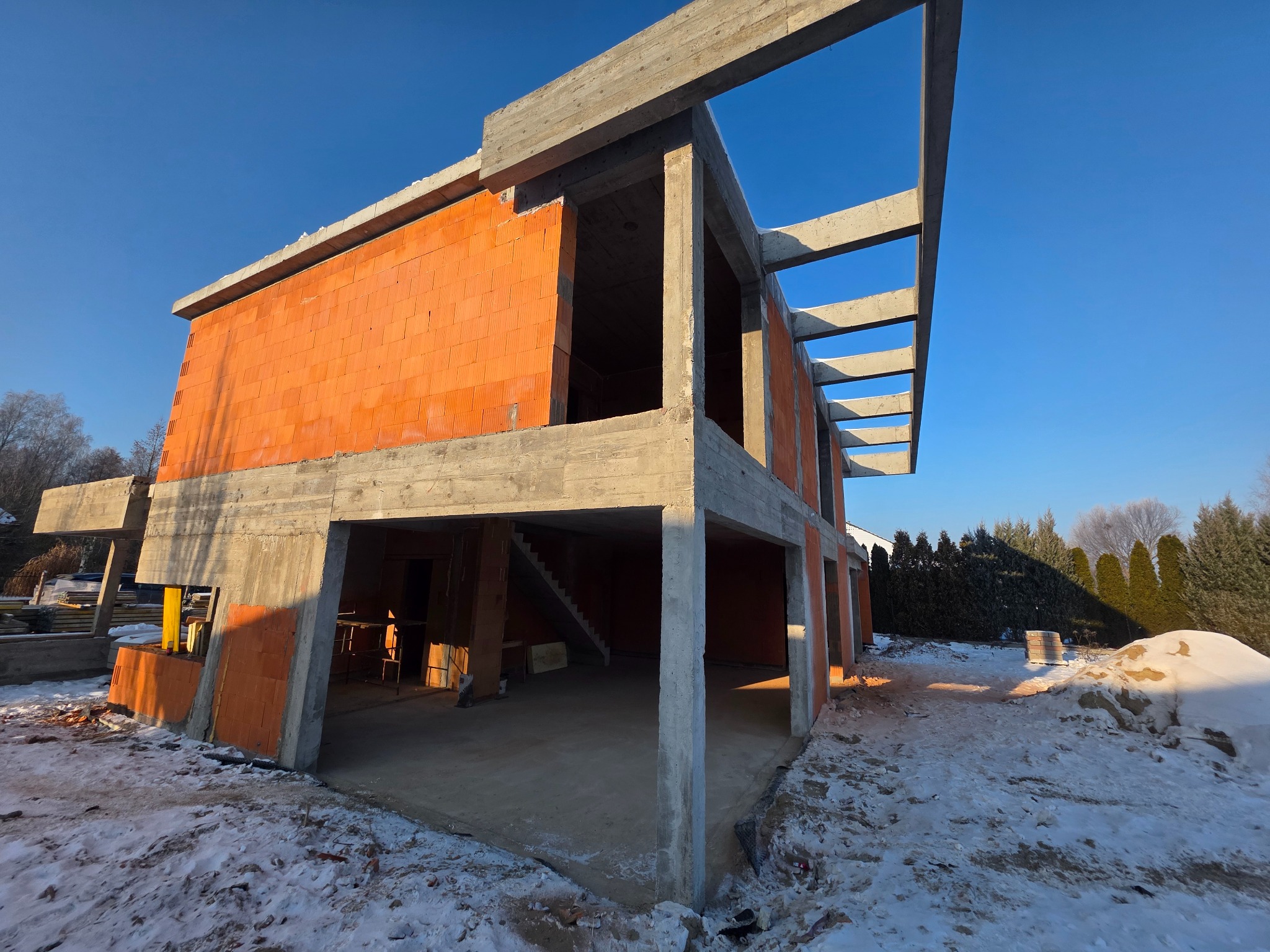 Surowy stan domu z czerwonej cegły i betonu w zimowej scenerii. Widoczne elementy konstrukcyjne i częściowo ośnieżone otoczenie. Budowa w toku, błękitne niebo w tle.