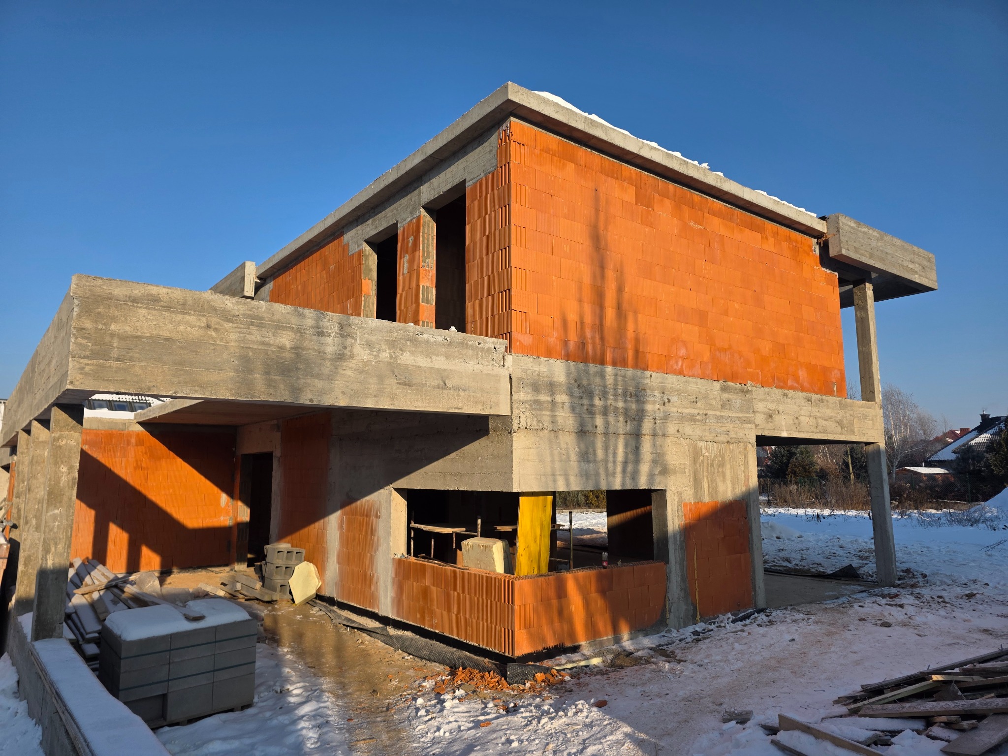 Surowy stan domu z czerwonej cegły i betonu w zimowej scenerii. Widoczne elementy konstrukcyjne i materiały budowlane. Budowa w toku, nieotynkowane ściany.