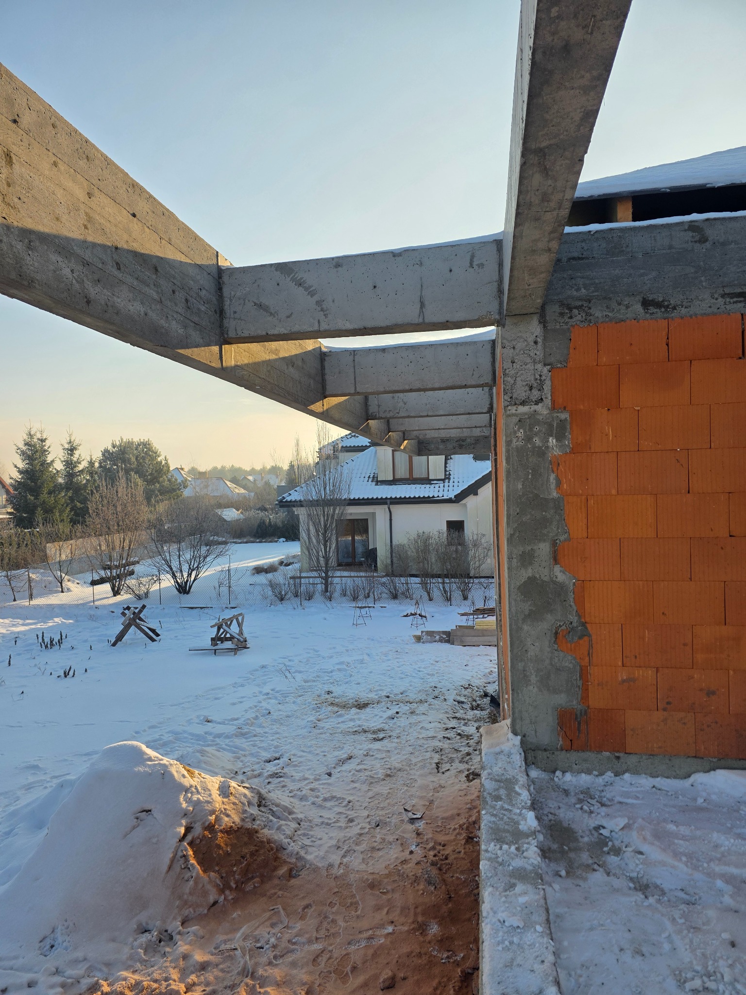Wznoszenie domu murowanego zimą: surowy stan z betonowymi belkami i fragmentem ściany z czerwonej cegły. W tle ośnieżony krajobraz i dom.