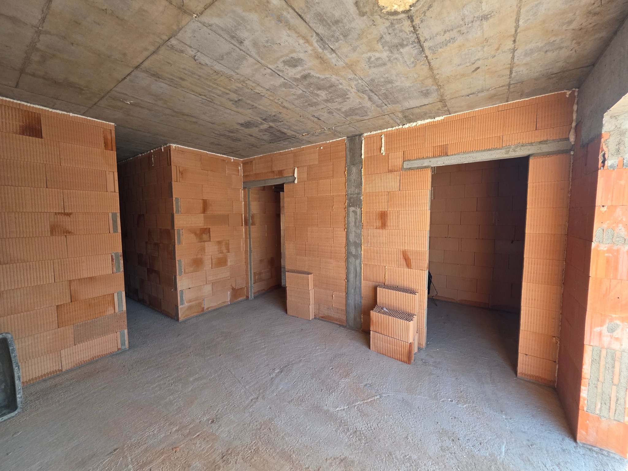 Wnętrze domu w trakcie budowy z czerwonej cegły, widoczne surowe ściany z otworami drzwiowymi i betonowy sufit. Na podłodze leżą cegły.