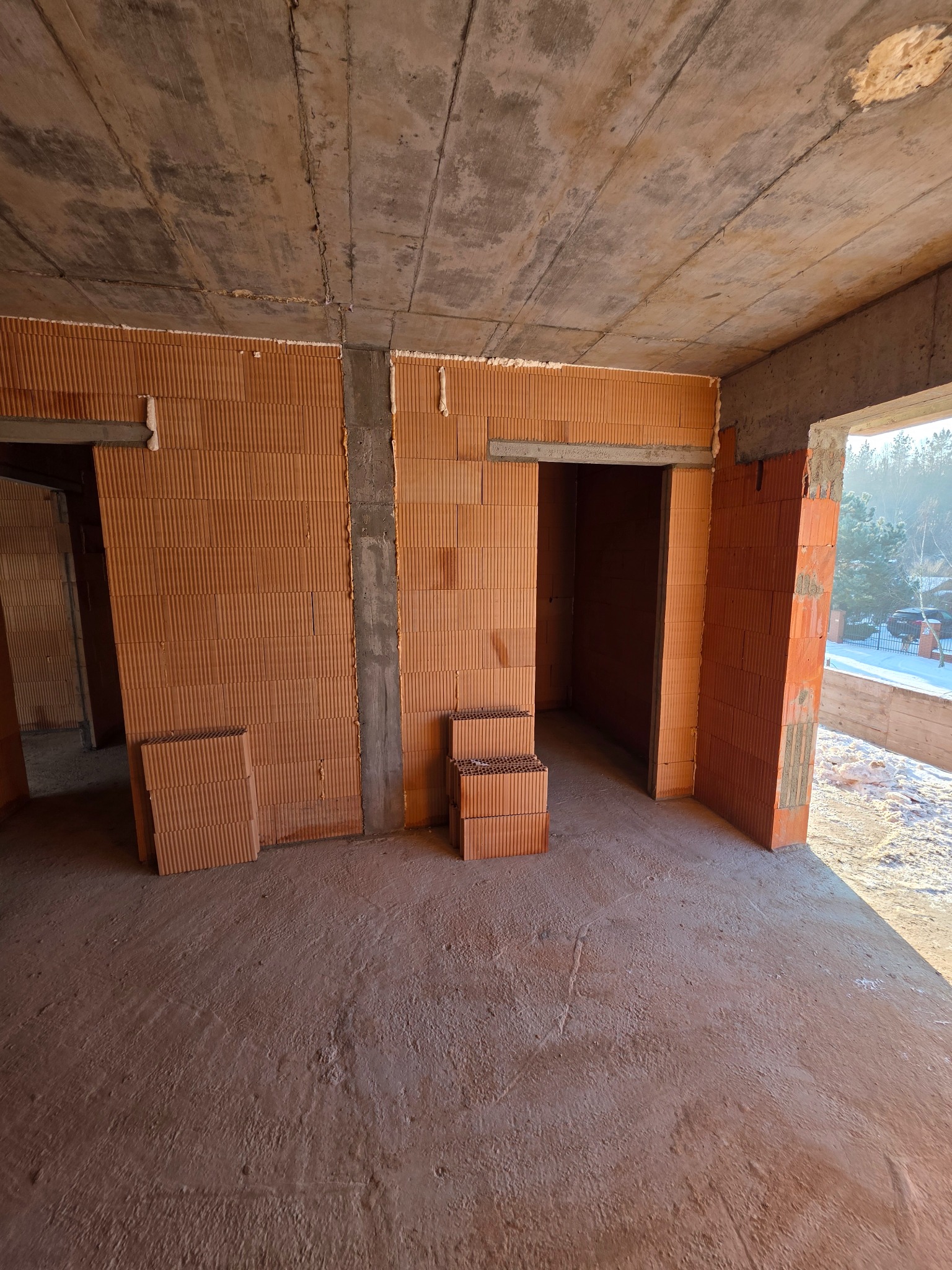 Wnętrze domu w trakcie budowy: ściany z czerwonej cegły, surowy betonowy strop, widoczne otwory drzwiowe i okienne, przygotowane do dalszych prac wykończeniowych.