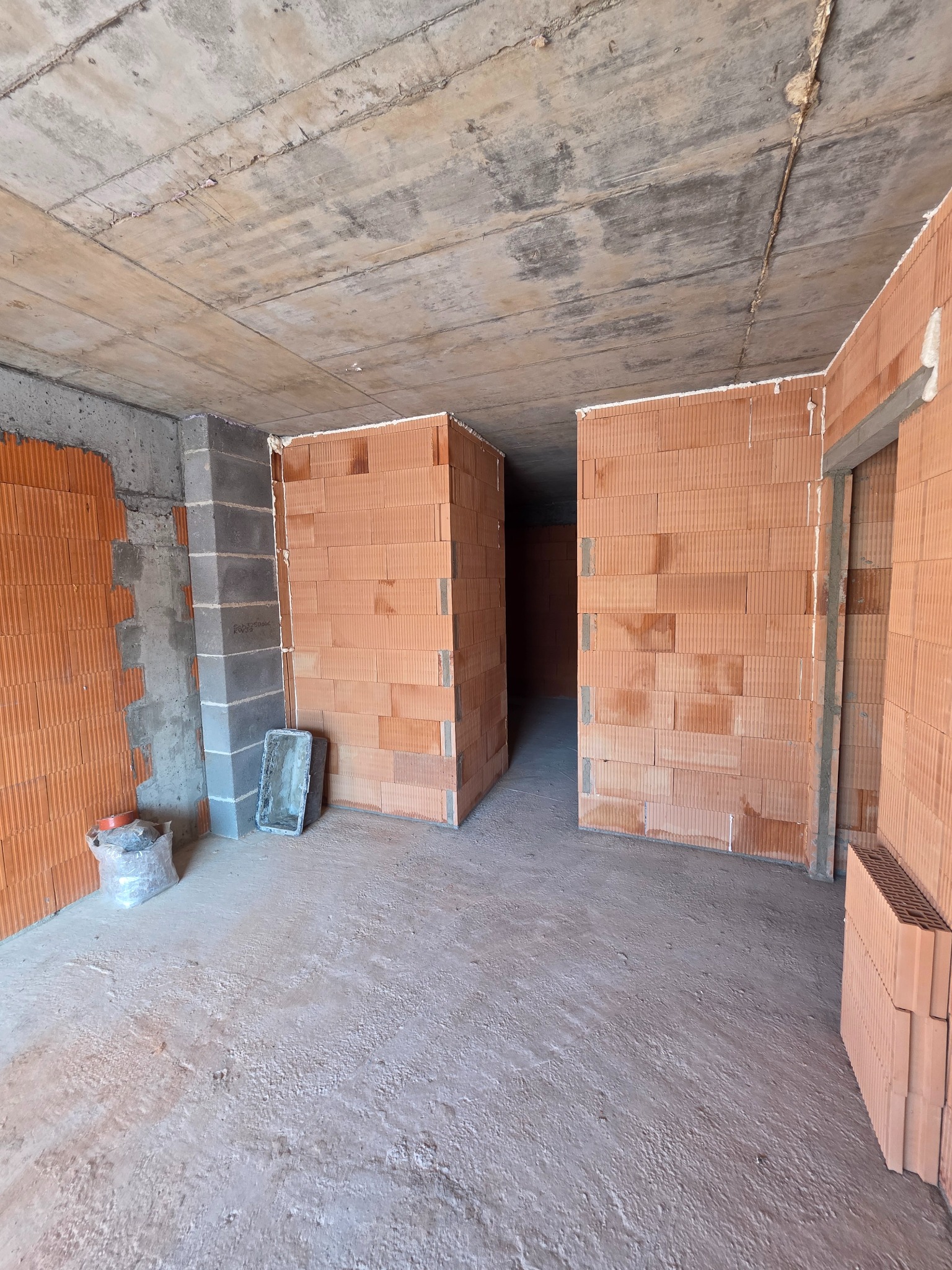 Wnętrze w trakcie budowy domu z czerwonej cegły, widoczne ściany działowe, surowy betonowy strop i narzędzia murarskie. Etap wznoszenia ścian.