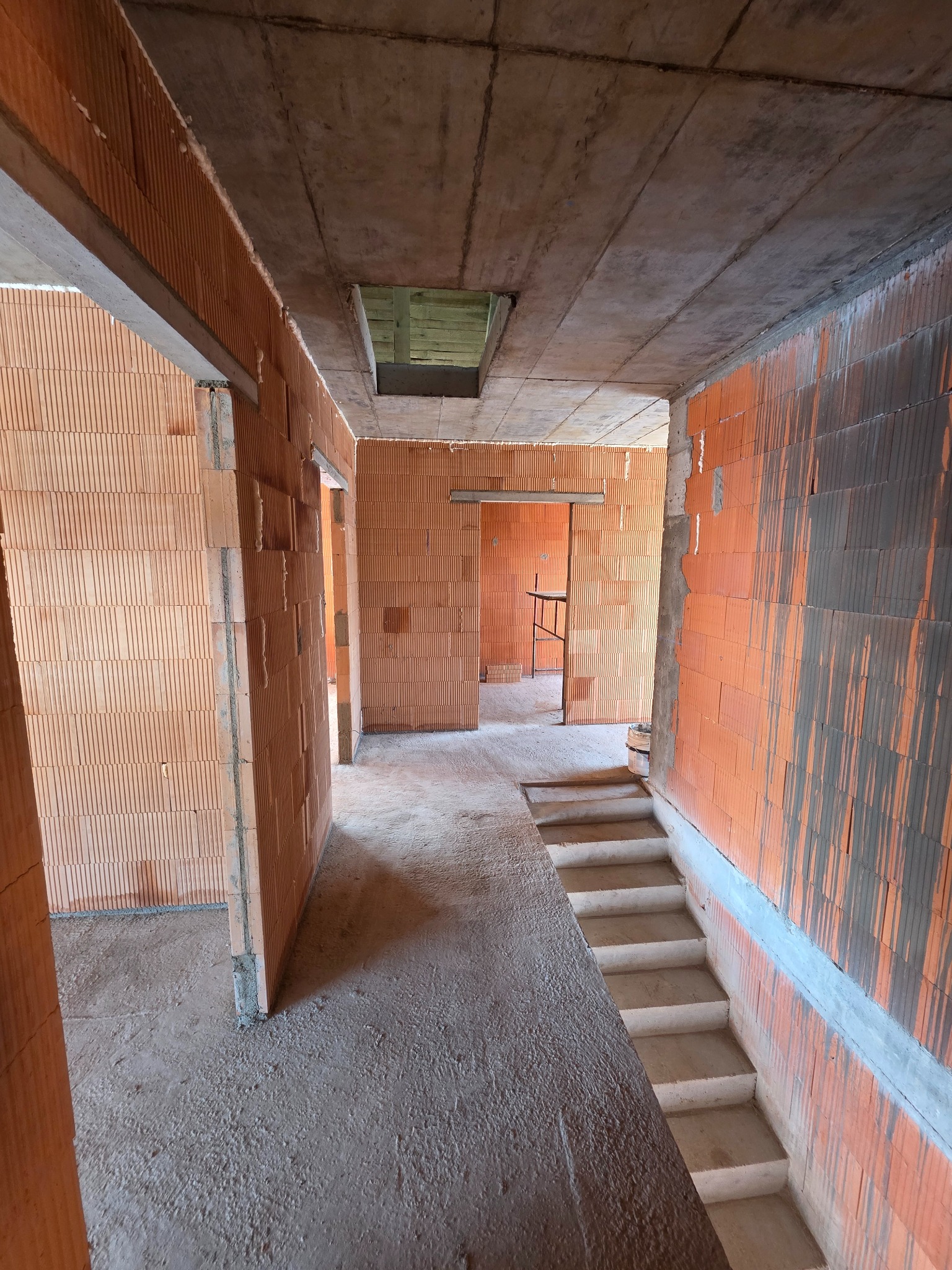 Wnętrze surowego domu w trakcie budowy: ściany z czerwonej cegły, betonowy strop, schody i widoczne przejścia do kolejnych pomieszczeń. W tle rusztowanie.