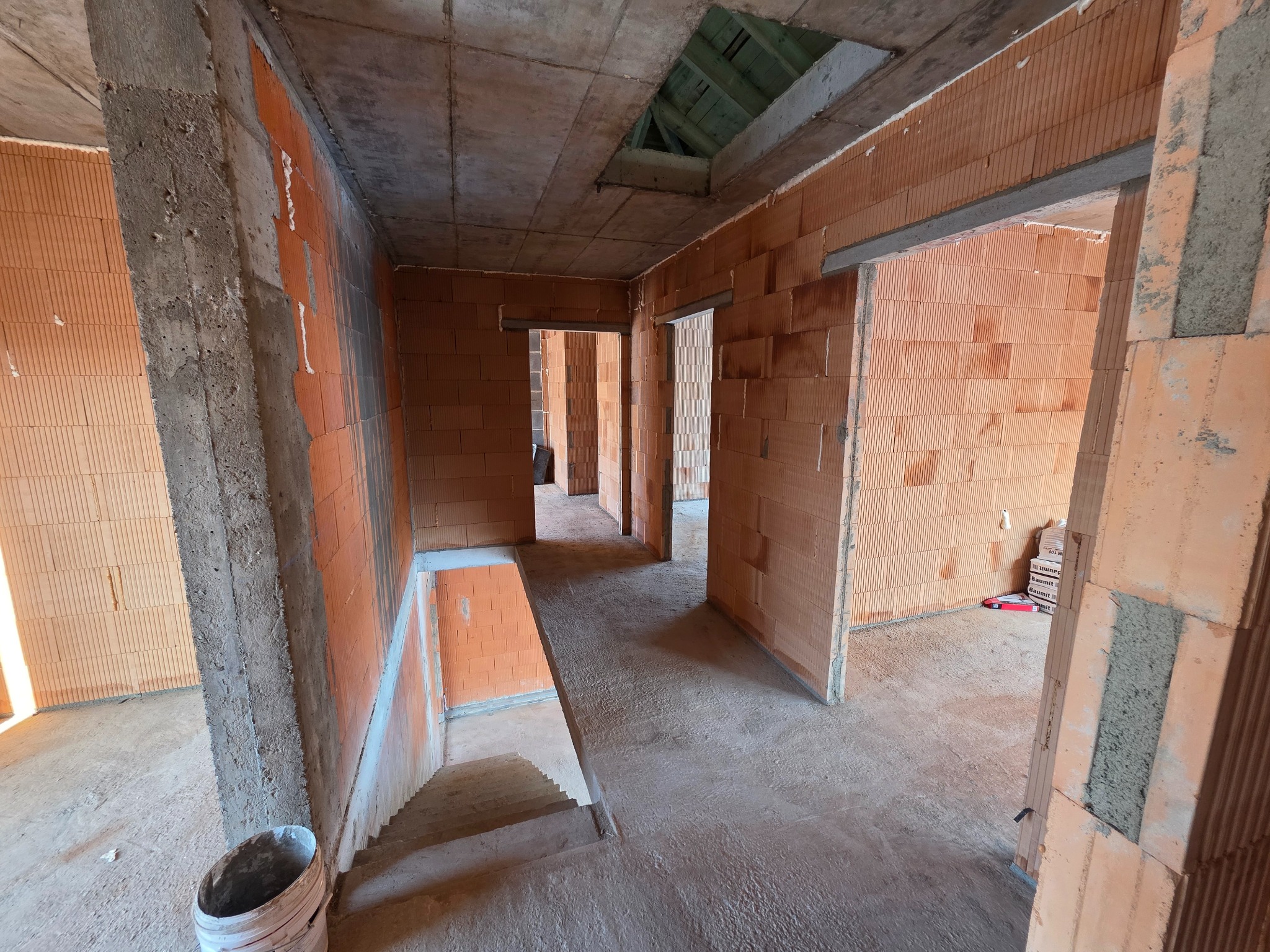 Wnętrze domu w trakcie budowy z czerwonej cegły, widoczne surowe ściany, betonowy strop z otworem i schody. Na podłodze widoczne ślady budowy, w tle otwarte przejścia.
