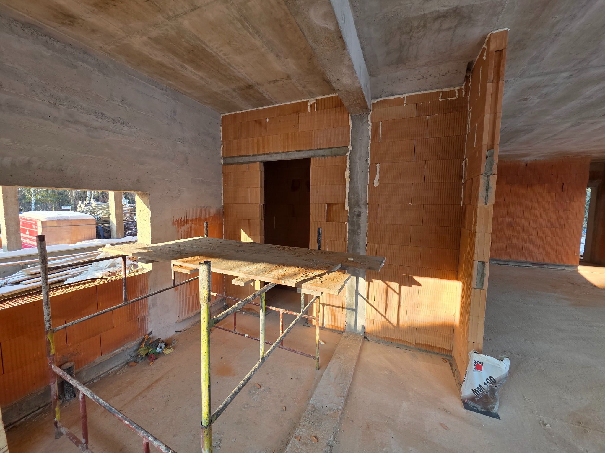 Wnętrze w trakcie budowy z ceglanymi ścianami, betonowym stropem i rusztowaniem. Widoczny worek zaprawy murarskiej. Surowy stan, przygotowanie do dalszych prac wykończeniowych.