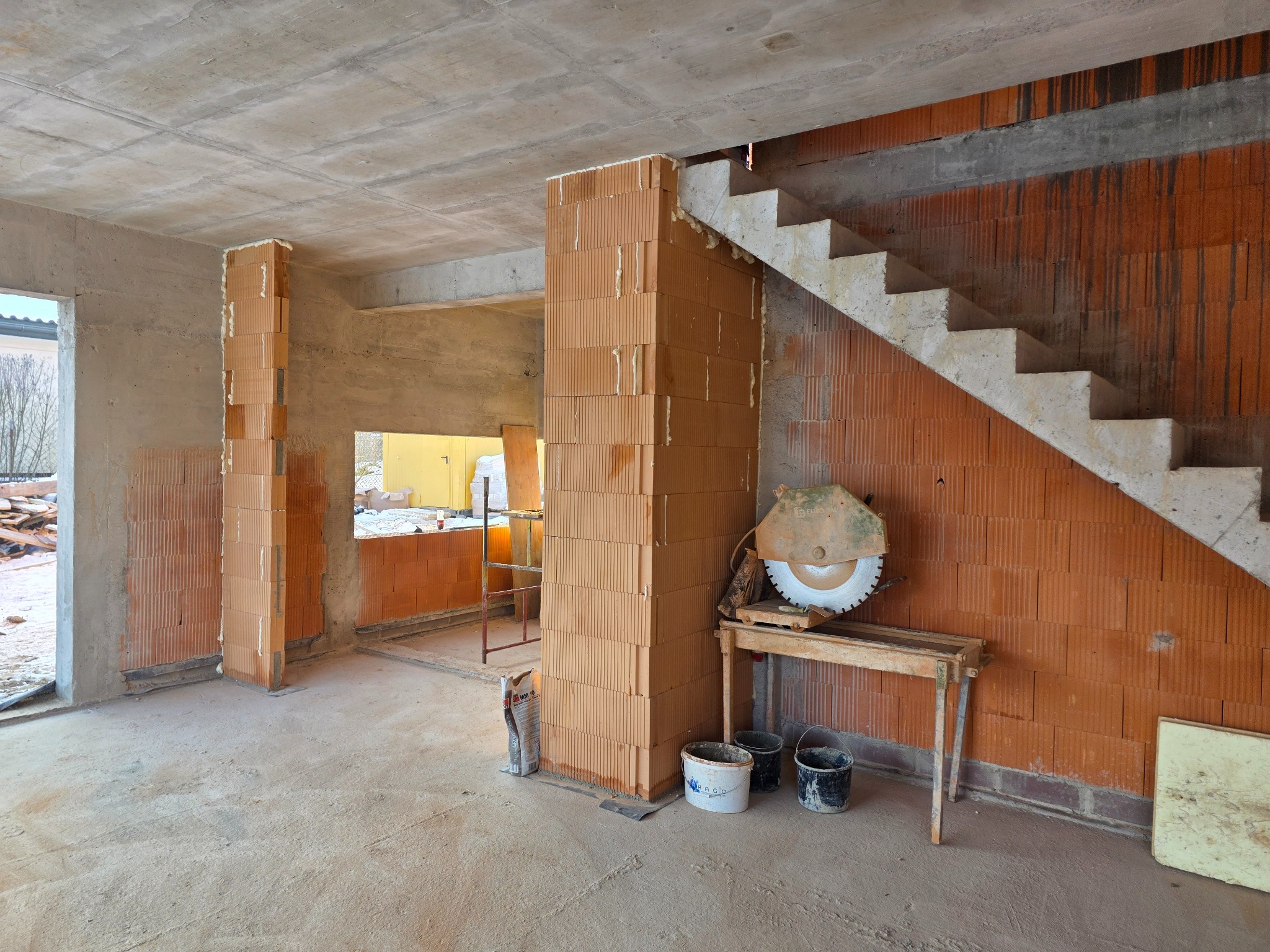 Wnętrze domu w trakcie budowy: ściany z cegły ceramicznej, surowy betonowy strop, schody, piła do cięcia cegieł na stole, worki z zaprawą. Widoczny etap murowania ścian.
