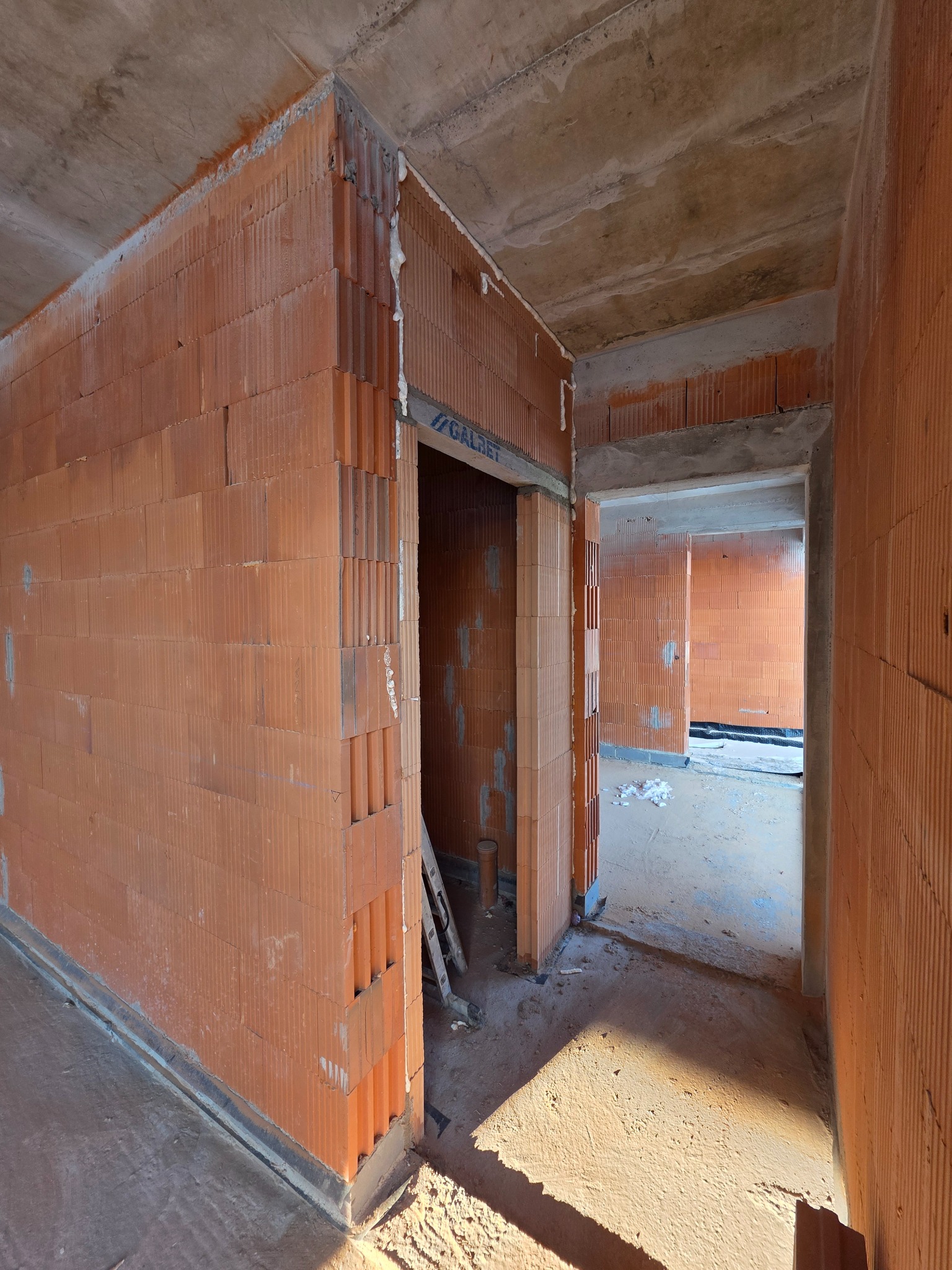 Wnętrze domu w trakcie budowy: ściany z czerwonej cegły, surowy betonowy strop, widoczne otwory drzwiowe i fragment drabiny. Stan surowy zamknięty.