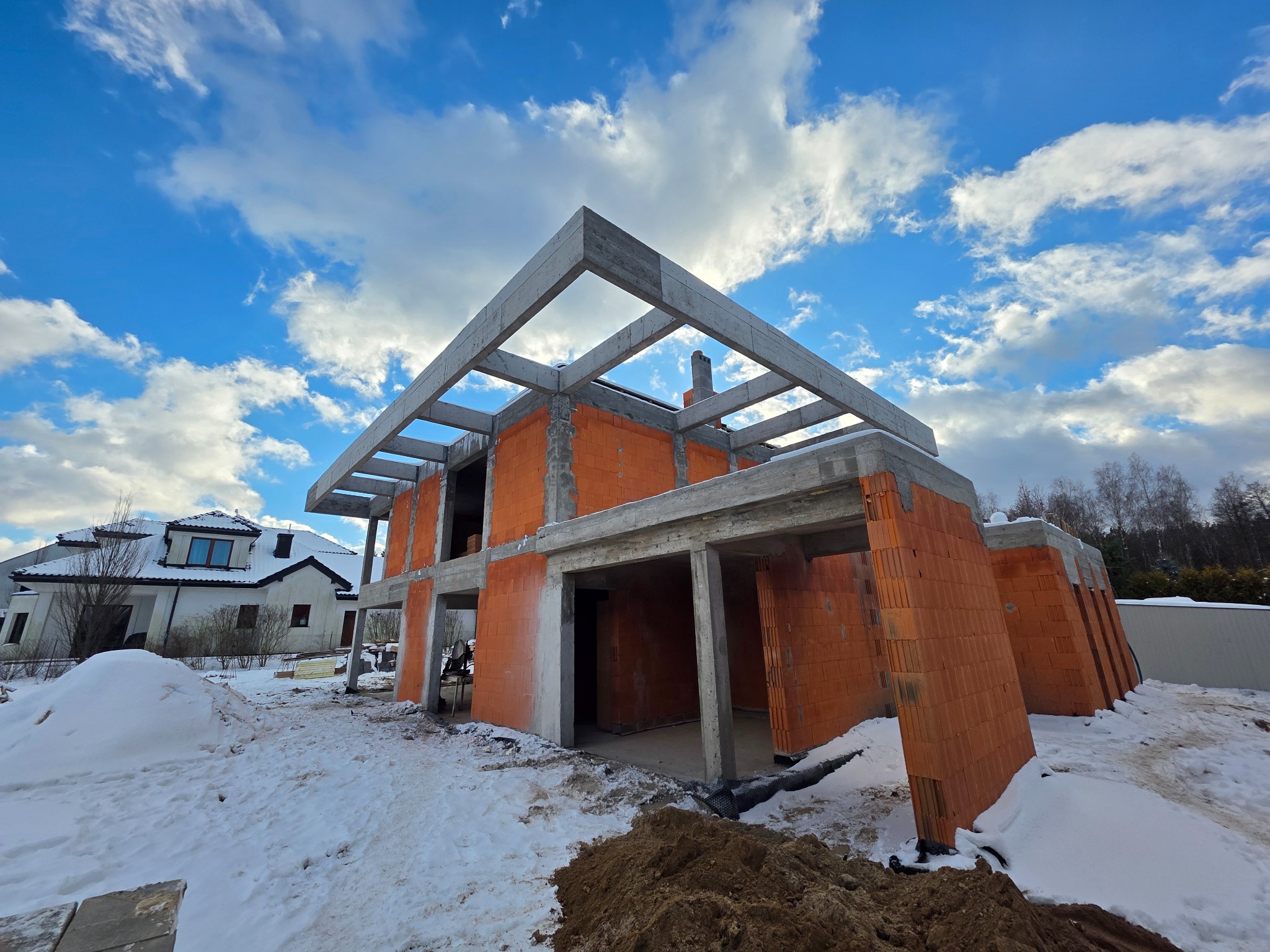 Surowy stan domu z czerwonej cegły i betonowych belek stropowych pod błękitnym niebem z chmurami. W tle widoczny inny dom i śnieg.