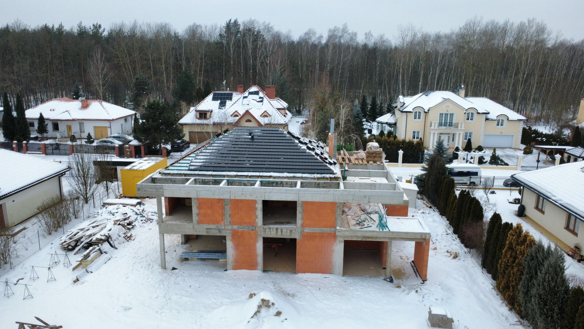 Budowa domu z czerwonej cegły w zimowej scenerii. Częściowo pokryty dach z czarnej dachówki. W tle inne domy i las. Widok z góry.