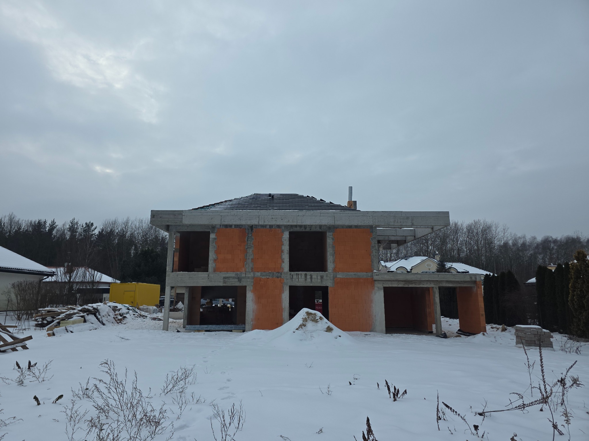 Surowy stan domu z czerwonej cegły w zimowej scenerii. Widoczne elementy konstrukcyjne i fundamenty. Na pierwszym planie śnieg i roślinność.