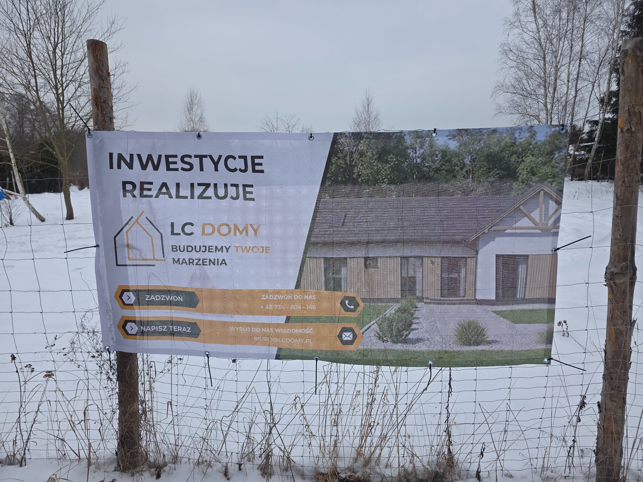Baner reklamowy firmy budowlanej LC Domy z wizualizacją domu murowanego, umieszczony na ogrodzeniu z siatki w zimowym krajobrazie. Widoczne dane kontaktowe i hasło: Budujemy Twoje Marzenia.