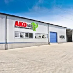 Budynek hali przemysłowej z szarej płyty warstwowej z niebieskimi akcentami, widoczne okna i brama wjazdowa, na ścianie logo firmy AKO ARES z zielonym elementem graficznym.