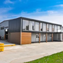 Nowoczesna hala produkcyjna z częścią biurową, dwukondygnacyjna, z elementami elewacji z płyt warstwowych i dużymi oknami, widok z zewnątrz, na pierwszym planie składowane elementy konstrukcyjne.