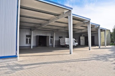 Hala magazynowa z otwartymi wjazdami, konstrukcja stalowa, ściany z płyt warstwowych, jasna kostka brukowa przed budynkiem, białe busy wewnątrz.