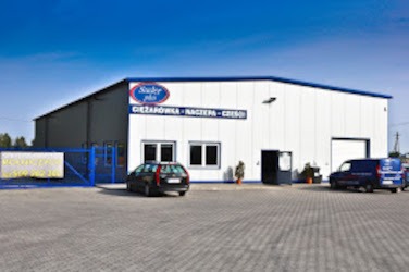 Biała hala produkcyjna z niebieskim logo firmy 'Staler Plus' nad wejściem, widoczny szary parking, czarny samochód osobowy i niebieski bus firmowy, błękitne niebo.