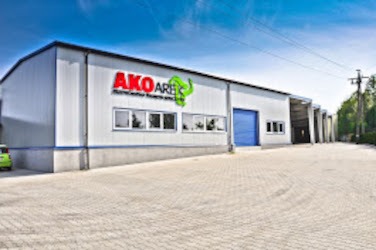 Budynek hali przemysłowej z szarej płyty warstwowej z niebieskimi akcentami, widoczne okna i brama wjazdowa, na ścianie logo firmy AKO ARES z zielonym elementem graficznym.