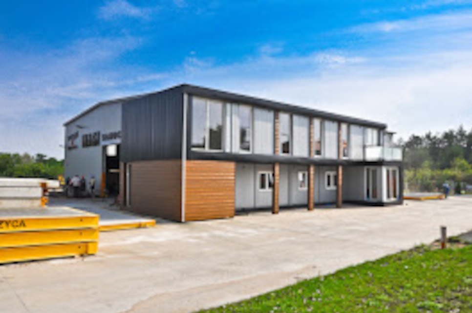 Nowoczesna hala produkcyjna z częścią biurową, dwukondygnacyjna, z elementami elewacji z płyt warstwowych i dużymi oknami, widok z zewnątrz, na pierwszym planie składowane elementy konstrukcyjne.