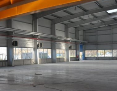 Wnętrze hali stalowej z betonową posadzką, widoczne okna z roletami, konstrukcja nośna w kolorze szarym i żółty suwnica pod stropem.