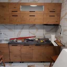 Mateusz nie - Montaż brązowych szafek kuchennych z poziomymi uchwytami na tle marmurowych płytek ściennych, widoczne narzędzia i akcesoria na blacie, w tle radio i okap kuchenny.