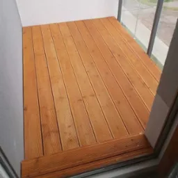 Wykończony balkon z drewnianą podłogą z desek kompozytowych w ciepłym odcieniu brązu, widok z góry na podłogę i fragment ściany z oknem.