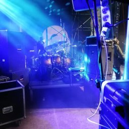 ESTUDIO WAV - Scena oświetlona niebieskimi reflektorami, z widoczną perkusją w tle i sprzętem nagłośnieniowym na pierwszym planie, w tym skrzynie transportowe i listwa zasilająca.