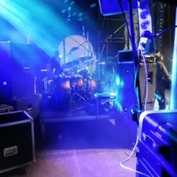 Scena oświetlona niebieskimi reflektorami, z widoczną perkusją w tle i sprzętem nagłośnieniowym na pierwszym planie, w tym skrzynie transportowe i listwa zasilająca.