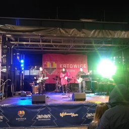ESTUDIO WAV - Zespół muzyczny występujący na scenie podczas Jarmarku Bożonarodzeniowego w Katowicach, widoczne instrumenty, oświetlenie sceniczne i banner reklamowy Jarmarku.