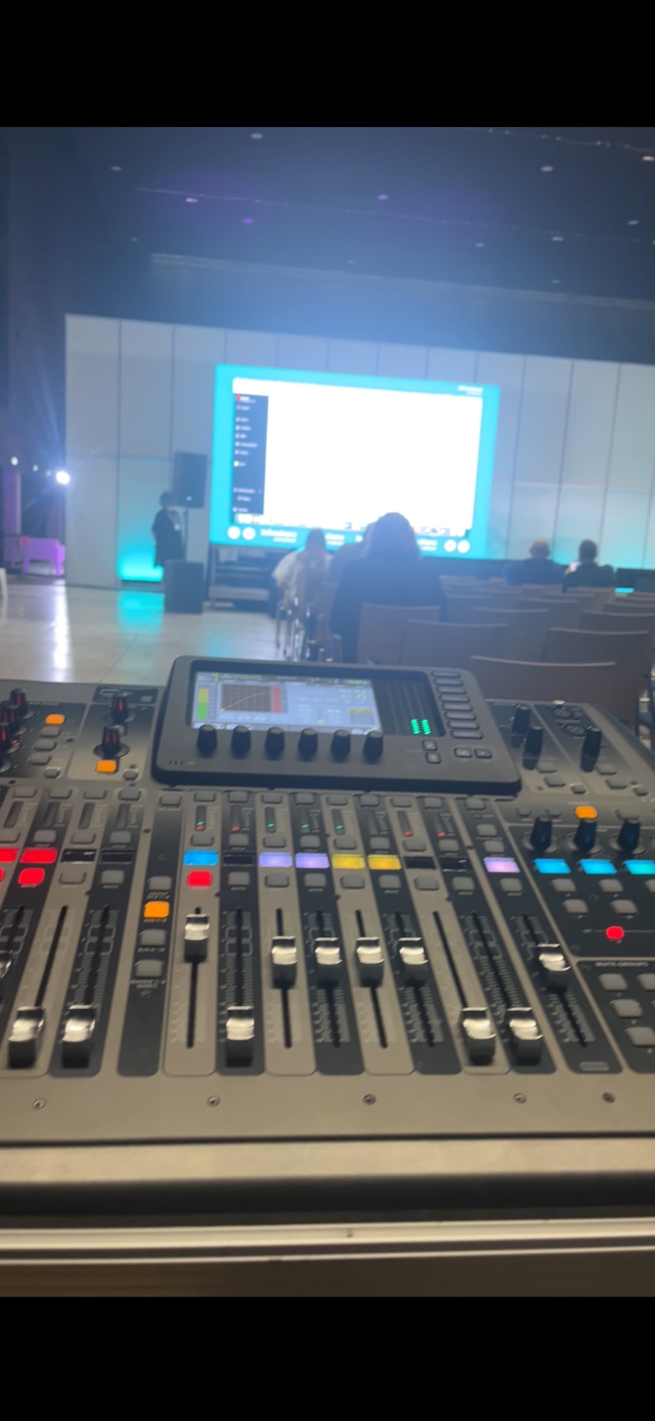 Profesjonalny mikser dźwięku z suwakami i pokrętłami, ustawiony przed sceną z publicznością i dużym ekranem.