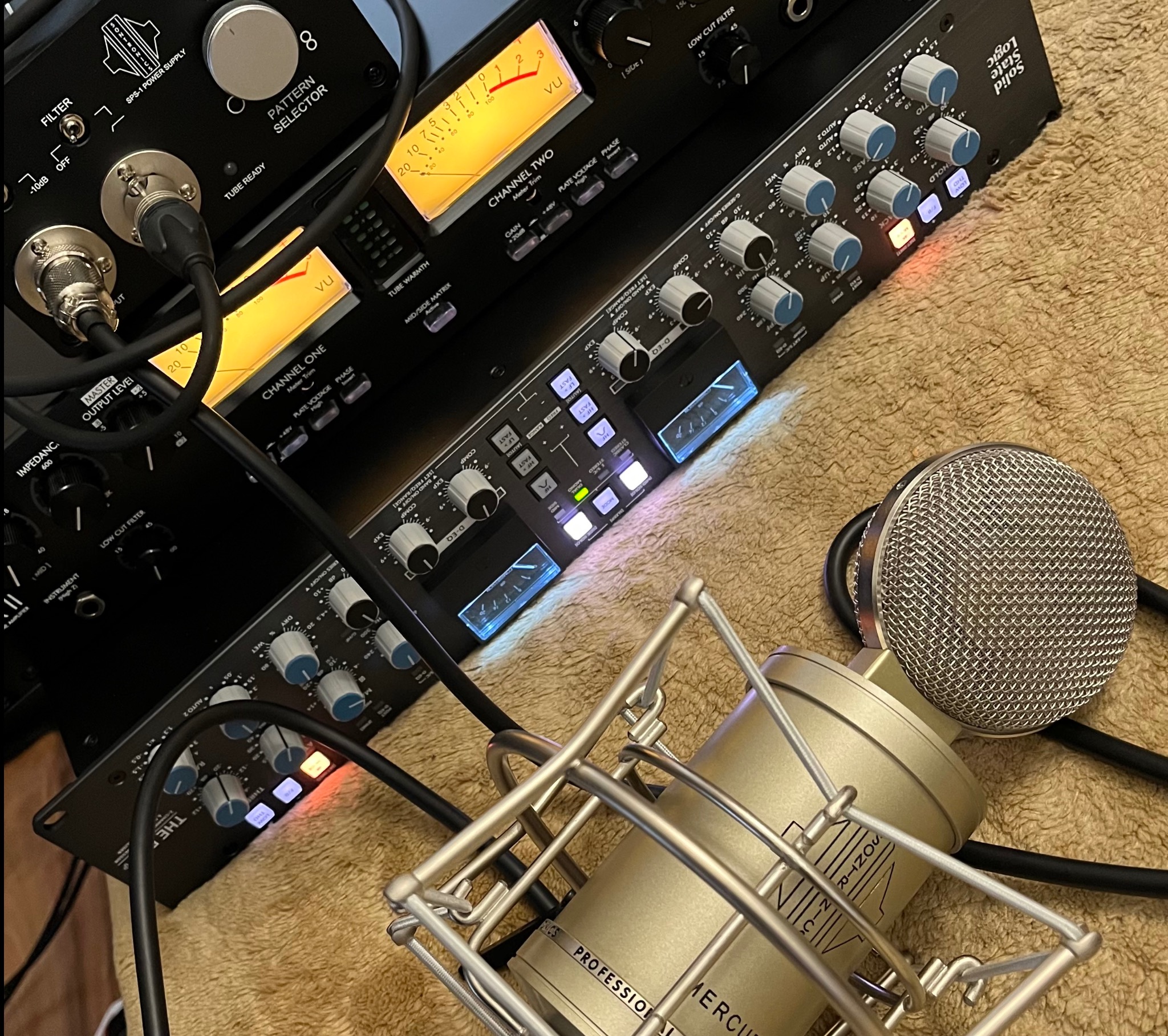 Mikrofon studyjny Mercury Professional SNICS na tle sprzętu audio: przedwzmacniacze, procesory dynamiki i equalizery. Widoczne analogowe wskaźniki VU i podświetlane przyciski.