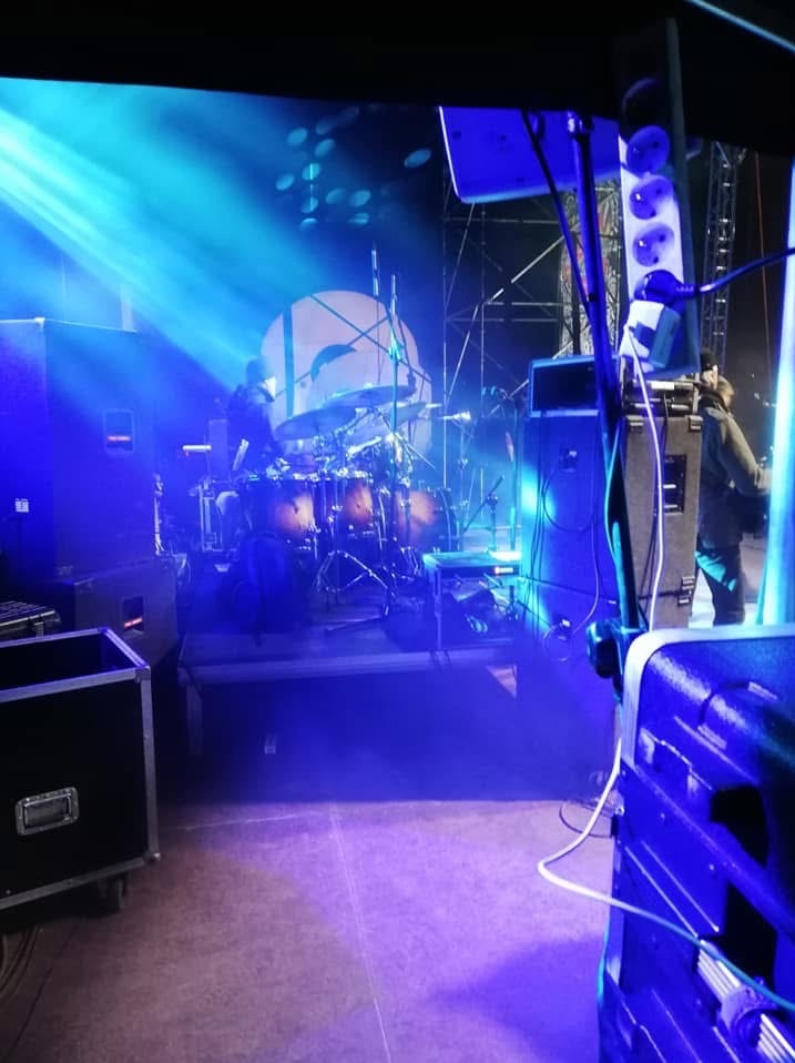 Scena oświetlona niebieskimi reflektorami, z widoczną perkusją w tle i sprzętem nagłośnieniowym na pierwszym planie, w tym skrzynie transportowe i listwa zasilająca.