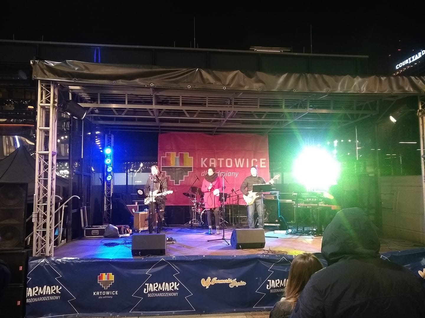 Zespół muzyczny występujący na scenie podczas Jarmarku Bożonarodzeniowego w Katowicach, widoczne instrumenty, oświetlenie sceniczne i banner reklamowy Jarmarku.