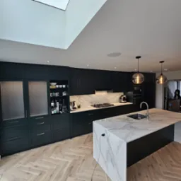 Nowoczesna kuchnia w odcieniach zieleni z marmurowym blatem wyspy, oświetlona wiszącymi lampami i światłem z okna dachowego, parkiet ułożony w jodełkę.