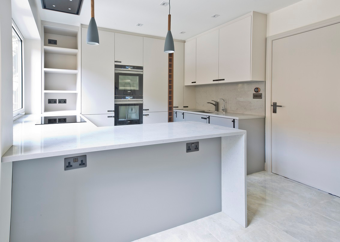 Nowoczesna, jasna kuchnia z wyspą, szarymi szafkami i białymi blatami, oświetlona wiszącymi lampami z drewnianymi elementami.