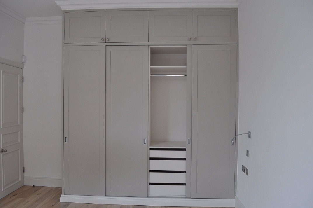 Szara szafa wnękowa z przesuwnymi drzwiami i górnymi szafkami, z widocznymi szufladami i drążkiem na ubrania w otwartej części, umieszczona w jasnym pomieszczeniu z drewnianą podłogą.