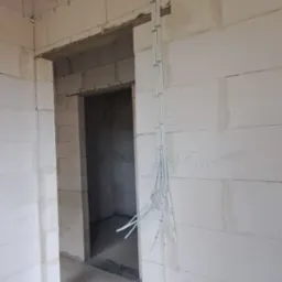 Wnętrze w stanie surowym z widocznymi przewodami elektrycznymi poprowadzonymi po ścianie z bloczków betonowych i wystającymi z sufitu, widok na przejście do kolejnego pomieszczenia.