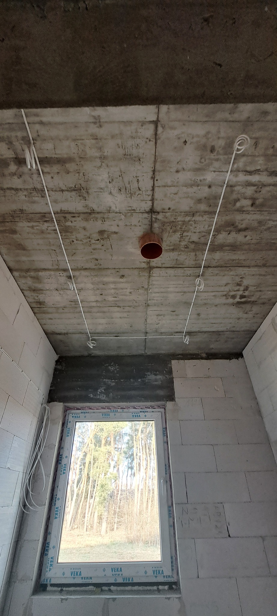 Surowy stan wnętrza z widocznymi przewodami elektrycznymi wyprowadzonymi z sufitu, pomarańczowa puszka instalacyjna na środku sufitu, okno z widokiem na las w tle.