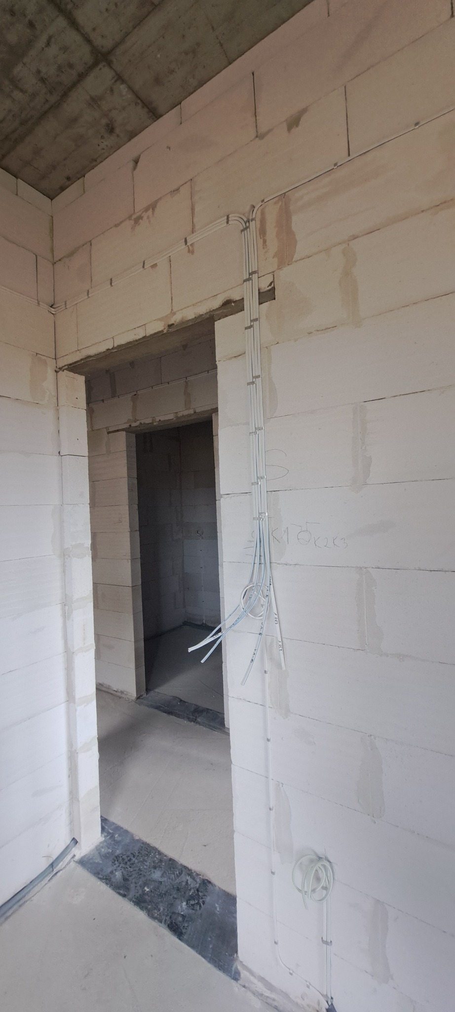 Wnętrze w stanie surowym z widocznymi przewodami elektrycznymi poprowadzonymi po ścianie z bloczków betonowych i wystającymi z sufitu, widok na przejście do kolejnego pomieszczenia.