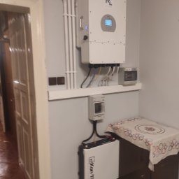 D&D Instal - Biały inwerter z wyświetlaczem, bateria oraz zabezpieczenia zamontowane na szarej ścianie wewnątrz pomieszczenia, instalacja elektryczna w białych rurkach.