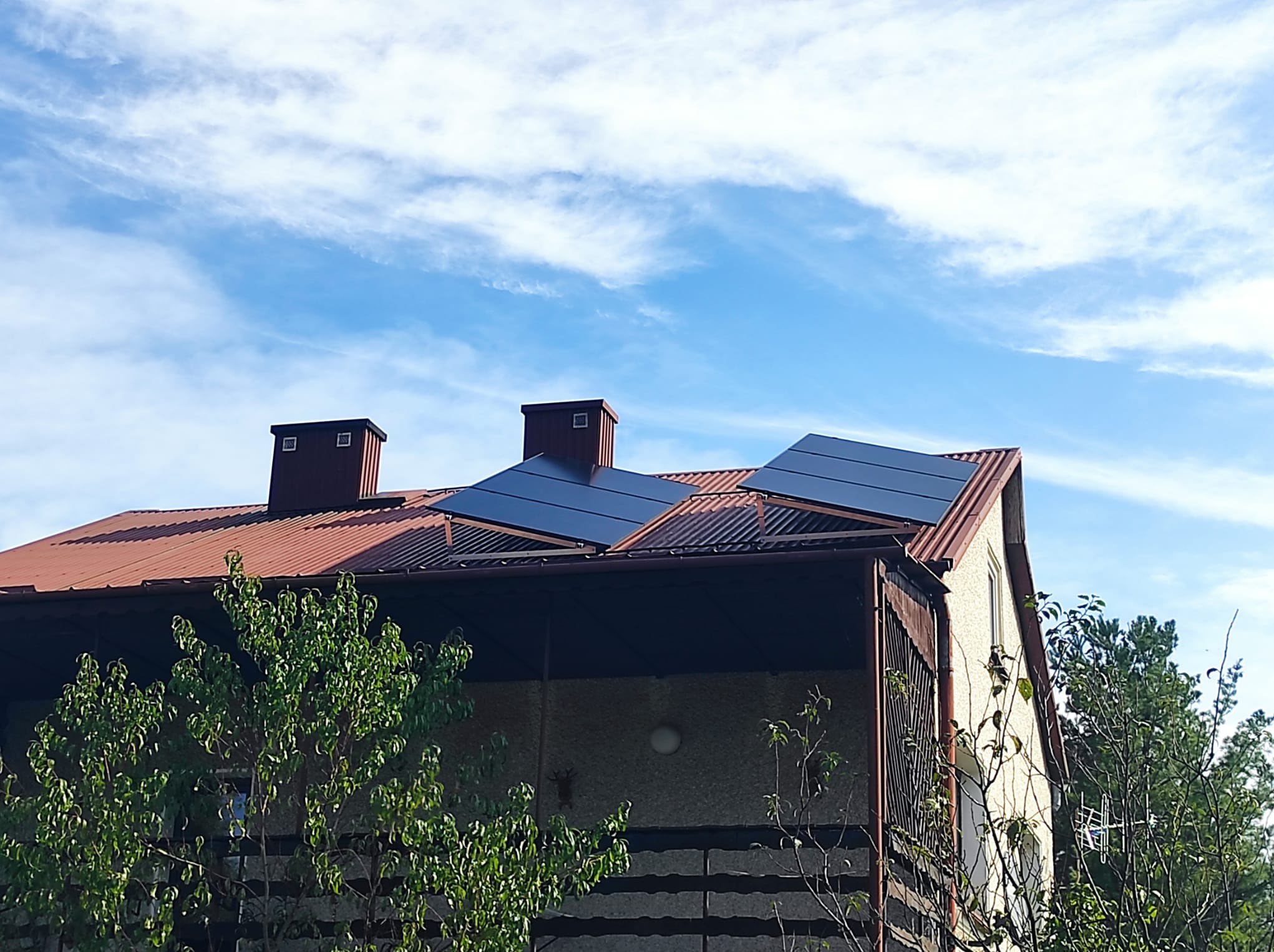 Panele słoneczne na dachu domu z blachy trapezowej, widok z dołu na tle błękitnego nieba z chmurami.