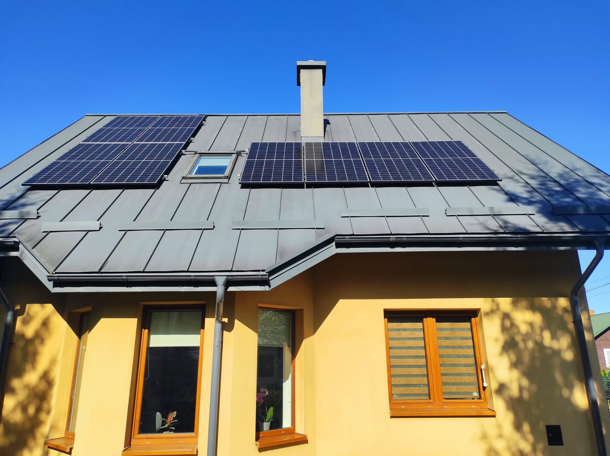 Panele słoneczne zamontowane na szarym, metalowym dachu domu z żółtą elewacją w słoneczny dzień. Widoczny komin i okno dachowe.