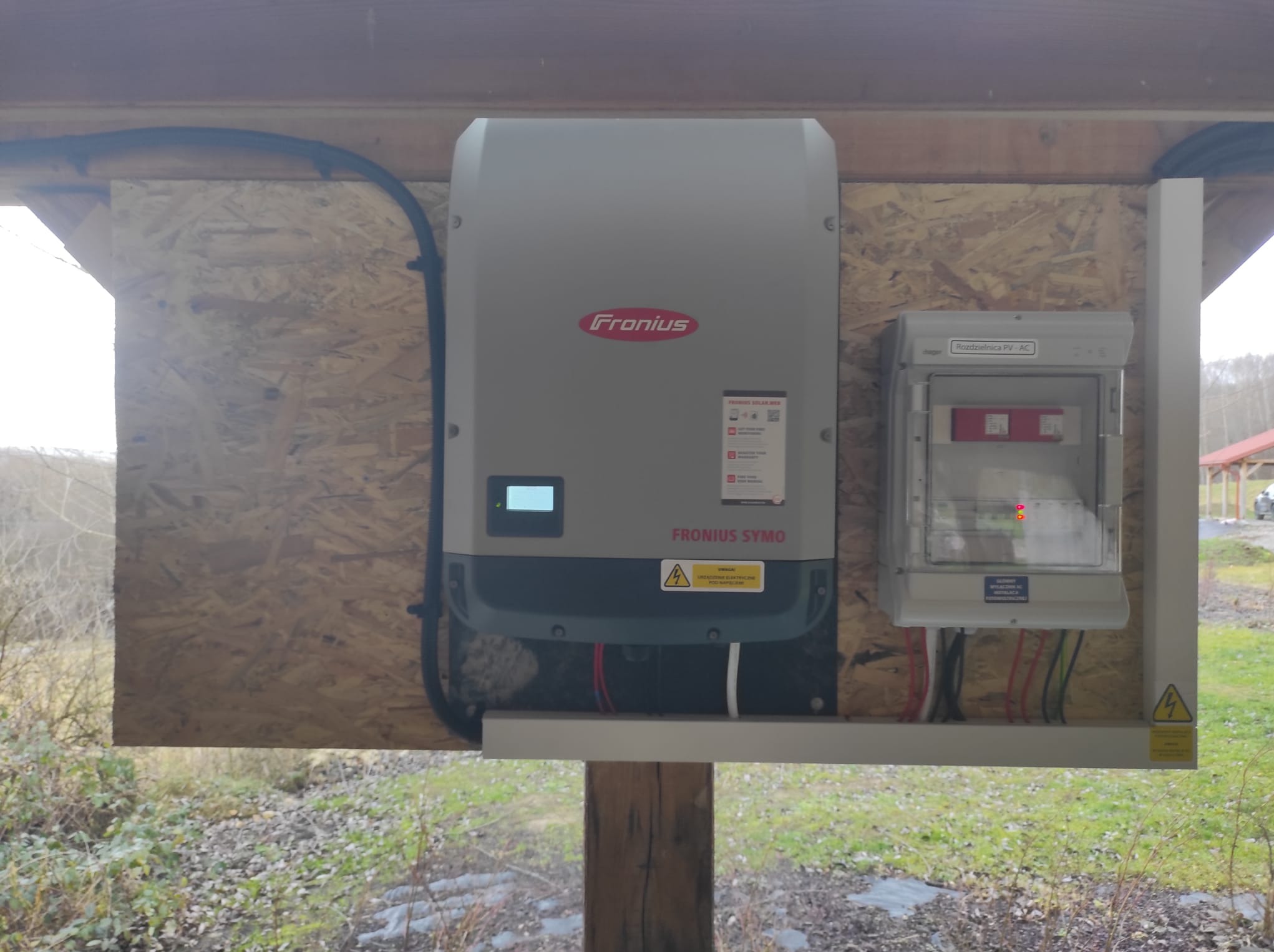 Szary falownik Fronius Symo i rozdzielnica PV-AC zamontowane na drewnianej konstrukcji na tle zieleni.