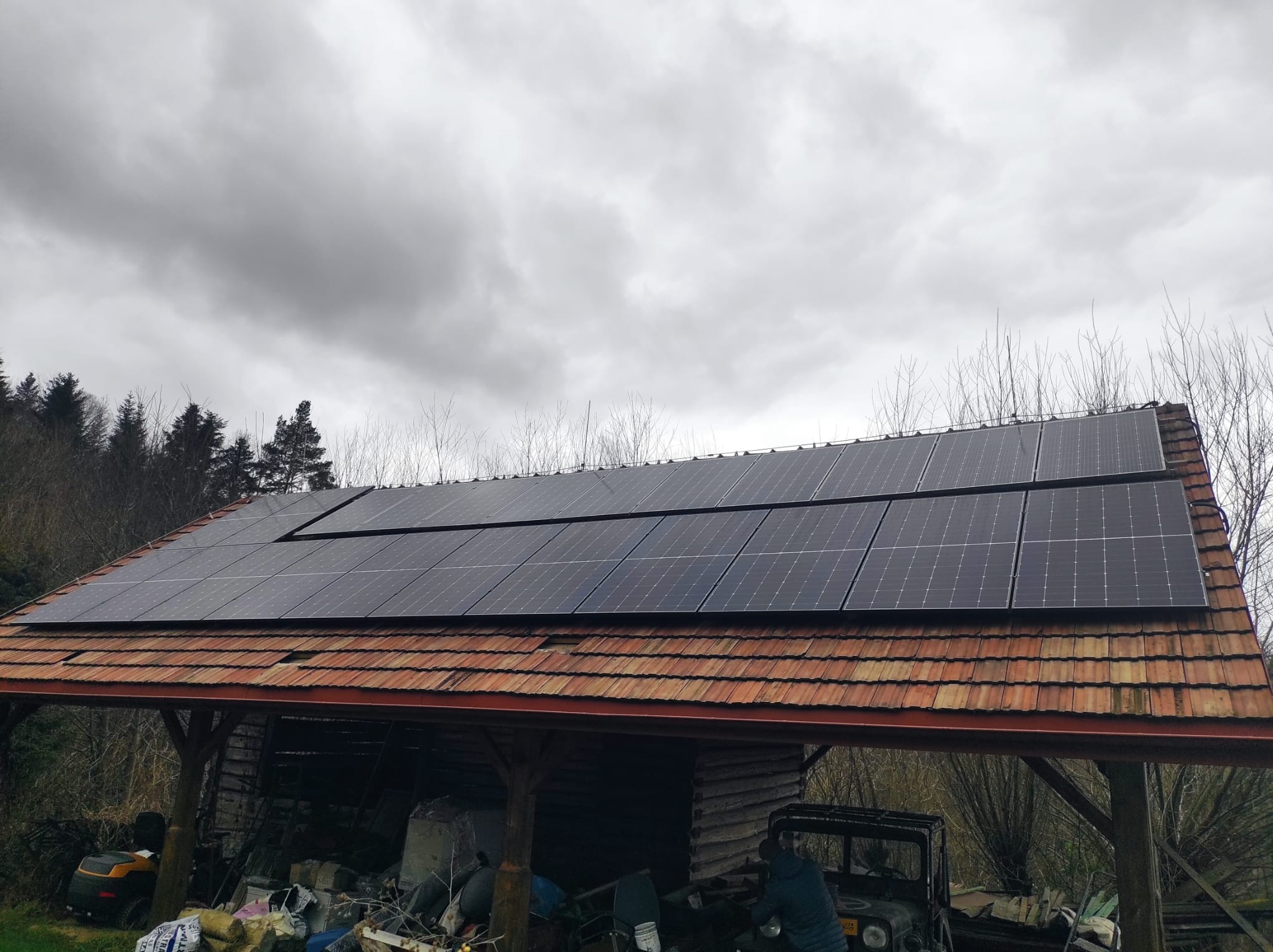 Panele słoneczne na dachu wiaty z czerwoną dachówką, pochmurne niebo w tle.
