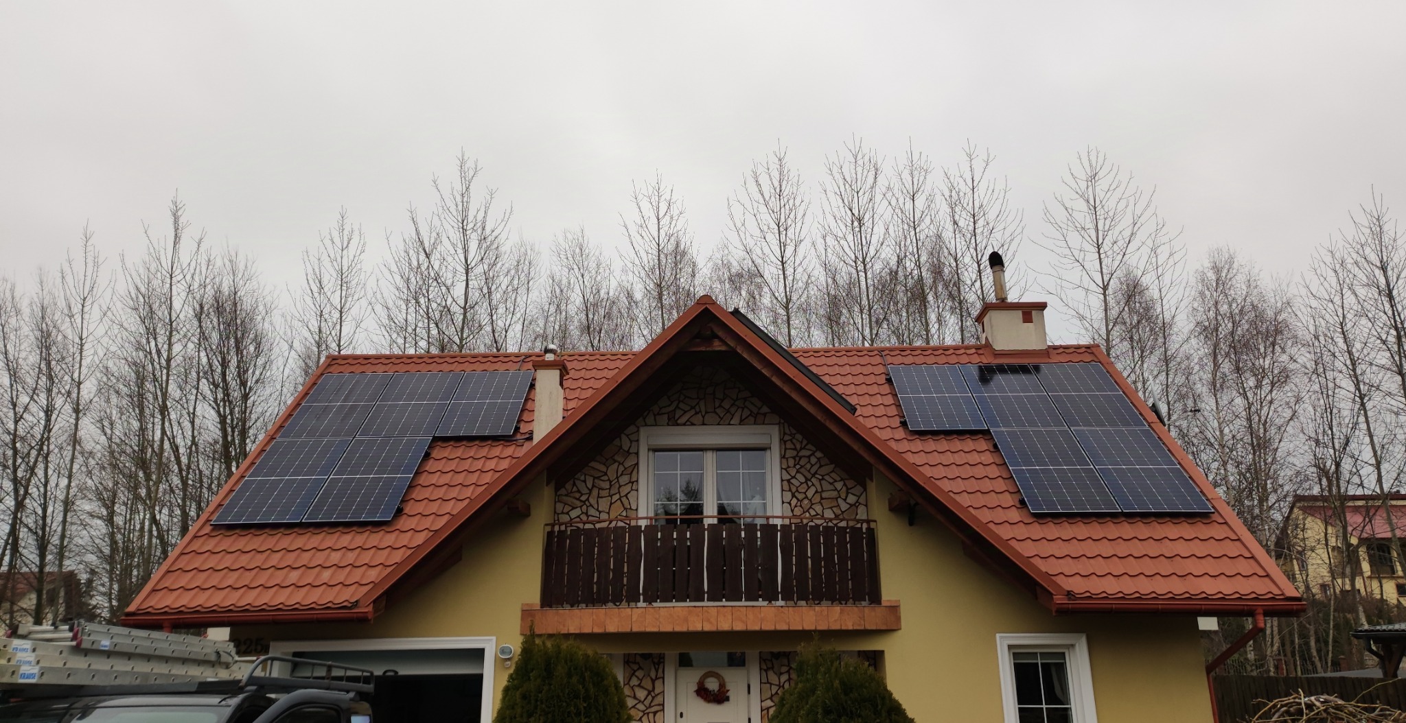 Panele fotowoltaiczne na dachu domu jednorodzinnego w Krasnem, widoczne drzewa w tle, dachówka ceramiczna w kolorze ceglastym.