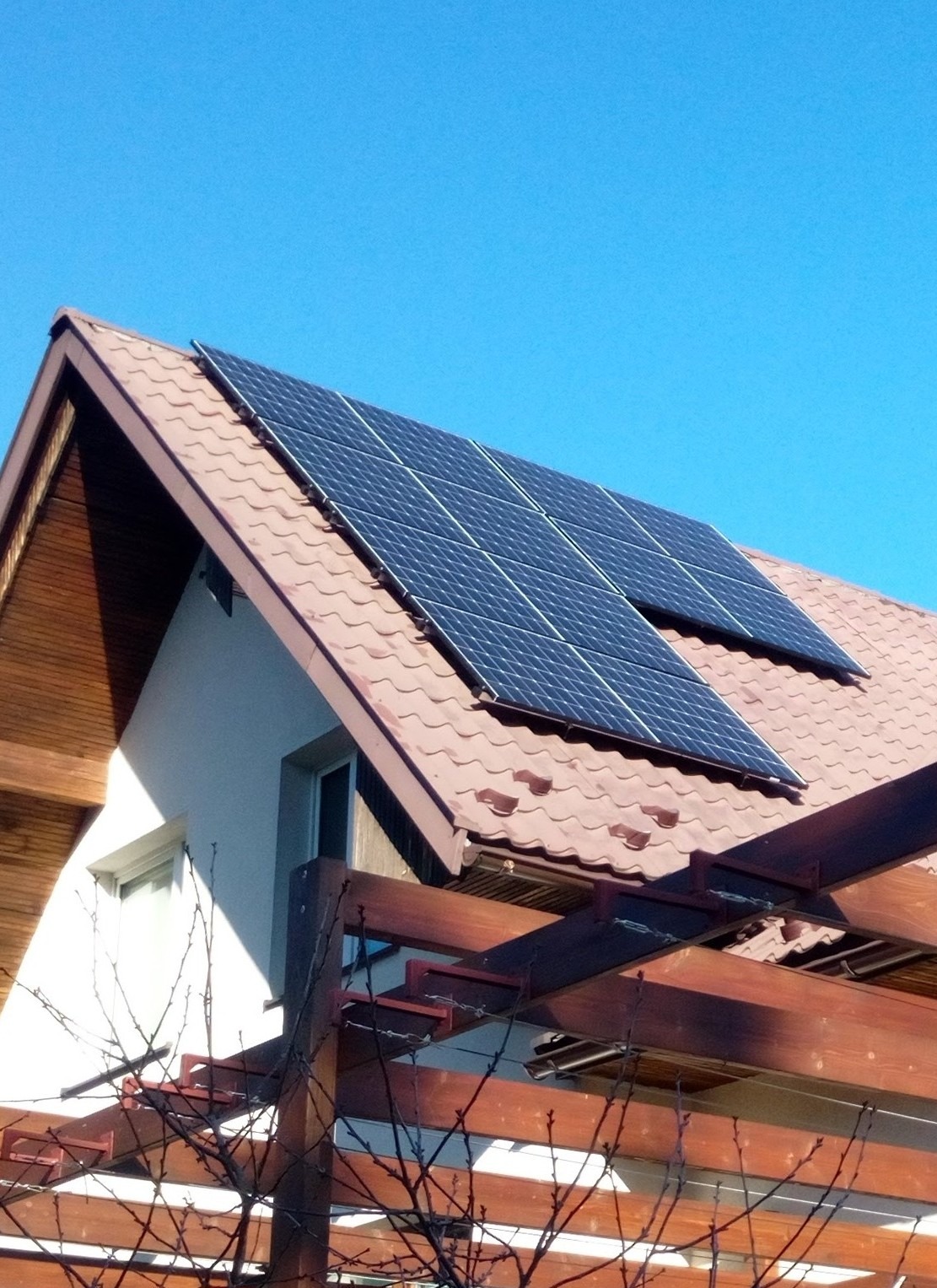 Dwa panele słoneczne na dachu domu z brązową dachówką, widoczne z perspektywy pergoli z ciemnego drewna na pierwszym planie, błękitne niebo w tle.