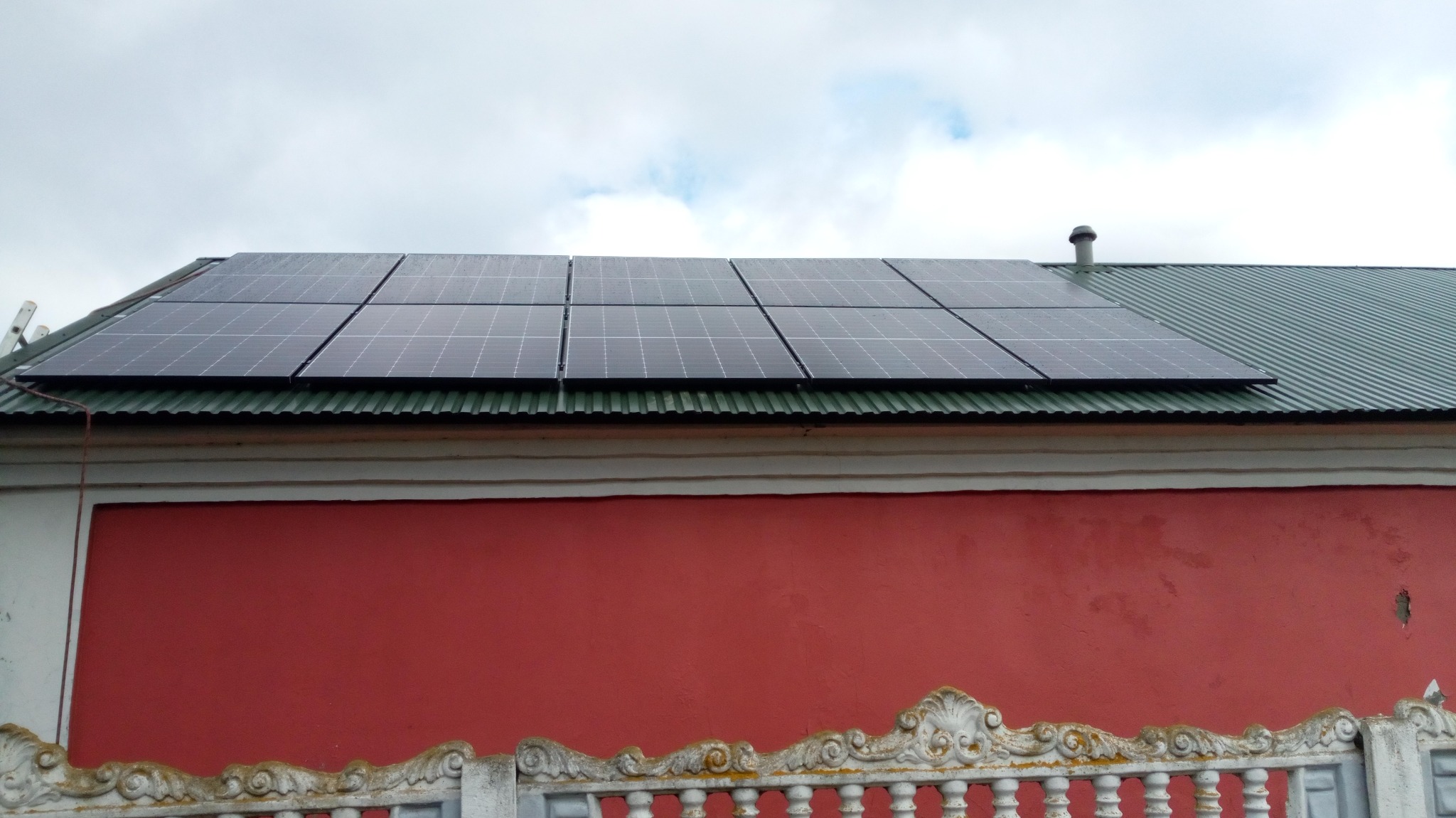 Panele słoneczne na blaszanym dachu budynku z czerwoną ścianą, widok z dołu, pochmurne niebo.